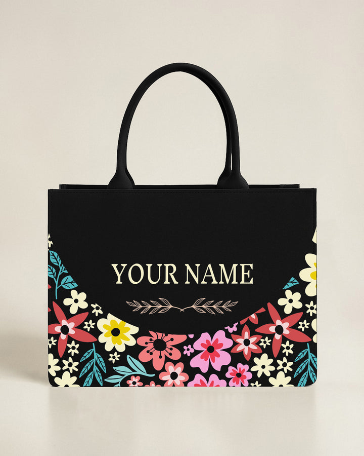 Black premium structured tote bag with a bottom crescent border of pink, red, and yellow flowers and a personalized name in serif font.