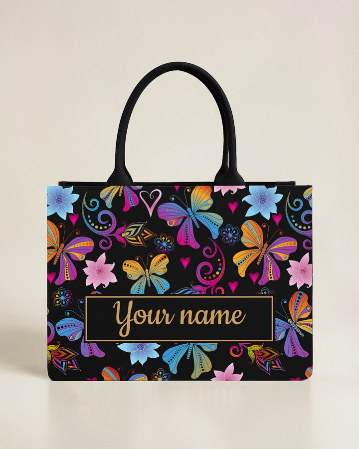 Black canvas tote bag with a lush bottom border of pink hibiscus flowers and tropical green palms, featuring a custom name in bold yellow font.