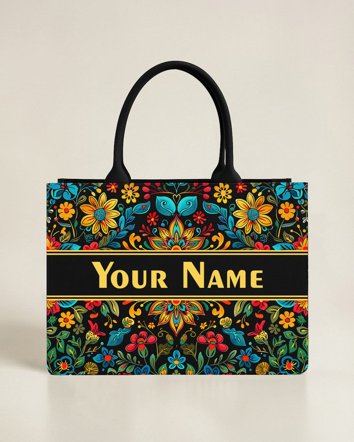 Black canvas tote bag with a geometric mosaic sun and mountain landscape in earthy red, blue, and gold tones, featuring a custom name in coral-pink font.