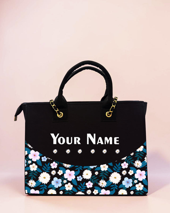 Personalized black tote bag with daisy floral artwork and custom name print displayed on a soft pastel background.