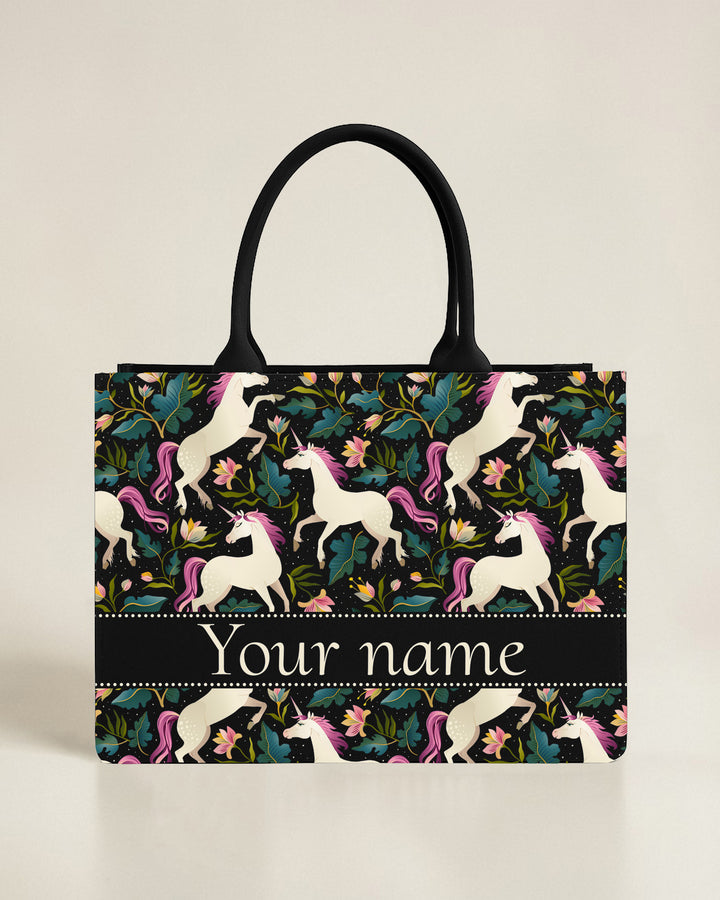 Black canvas tote bag featuring a vibrant teal hummingbird hovering over a border of pink flowers and green vines, with a custom name in bright lime-green font.