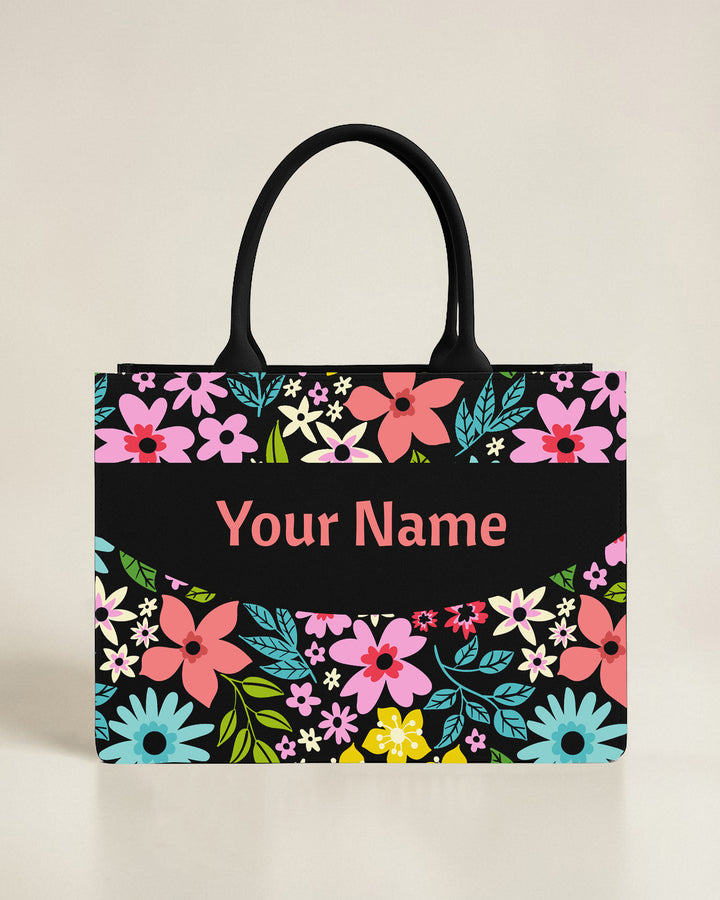 Personalized black tote bag featuring a full-frame border of oversized pink and blue flowers around a centered, curved name bar.