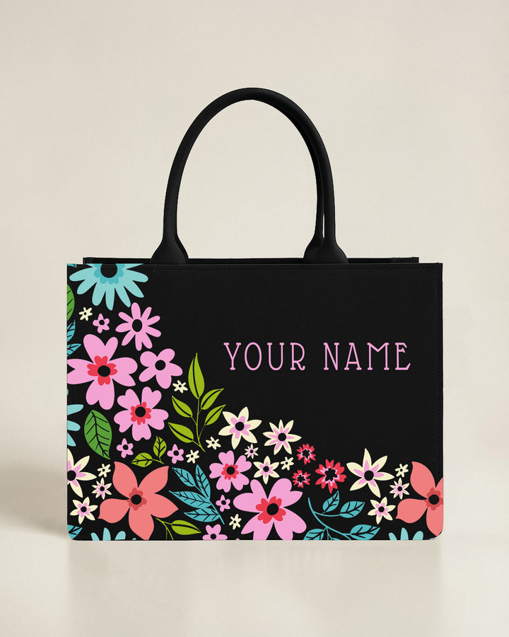 Black tote bag with a whimsical floral cluster growing from the bottom-left corner and a custom name in pink typewriter-style font.